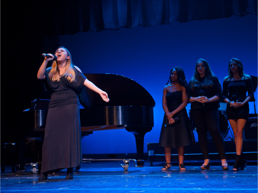 Female student singing on stage with backup singers and piano behind her