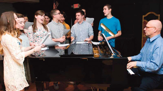 Students gathered around a piano singing while a teacher plays the piano