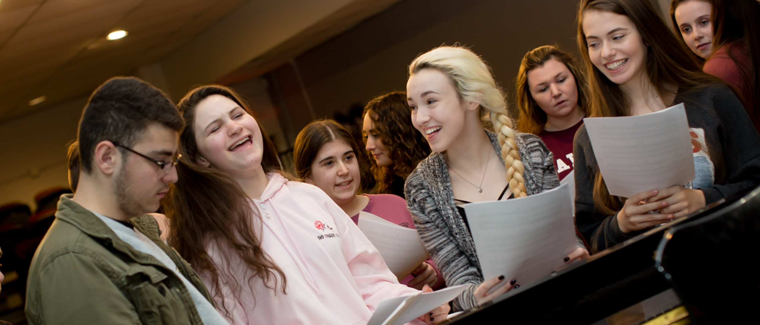 Students gathered around a piano with sheet music in their hands smiling and laughing