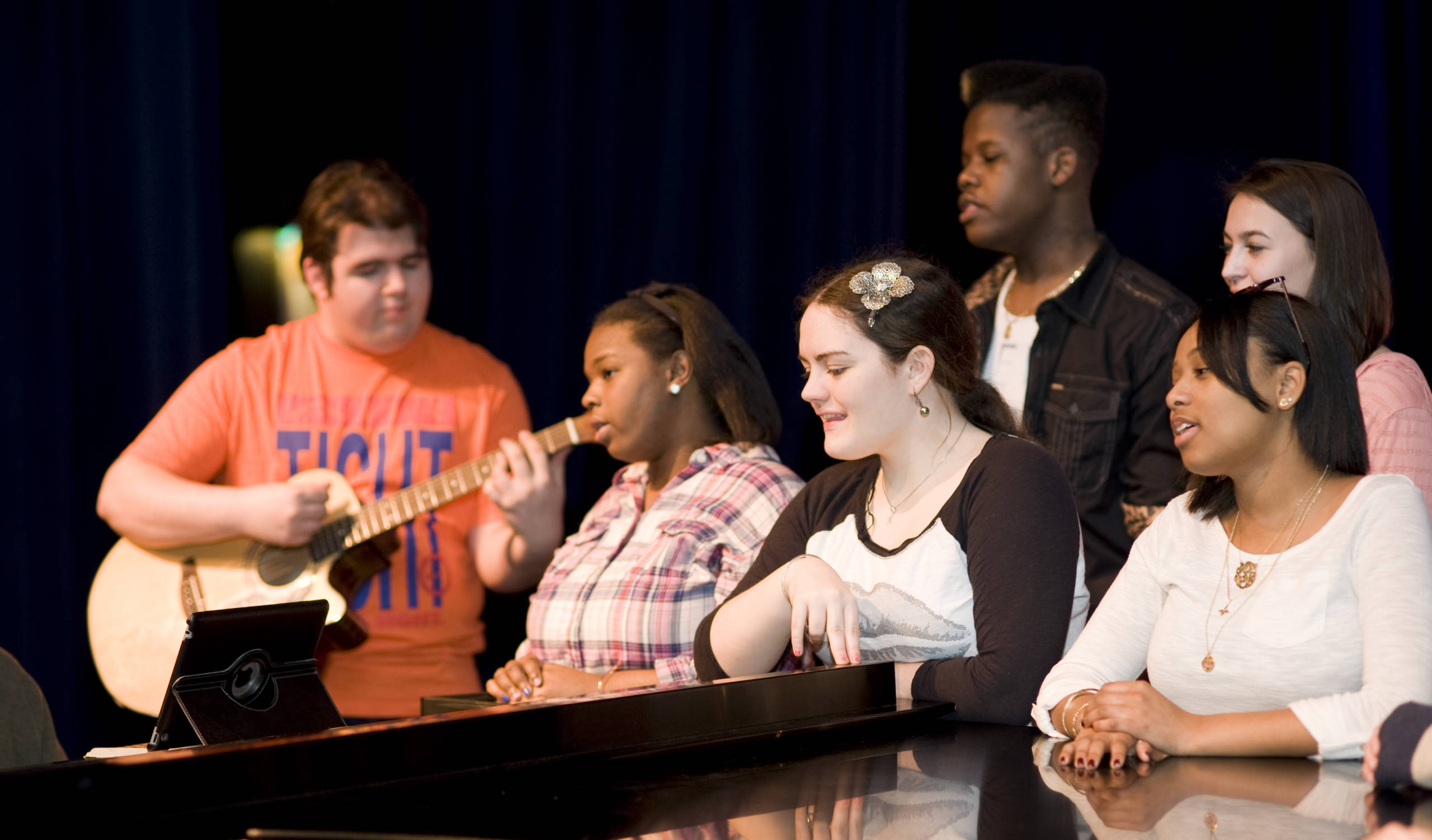 Students sing around a piano with a guitar accompanist